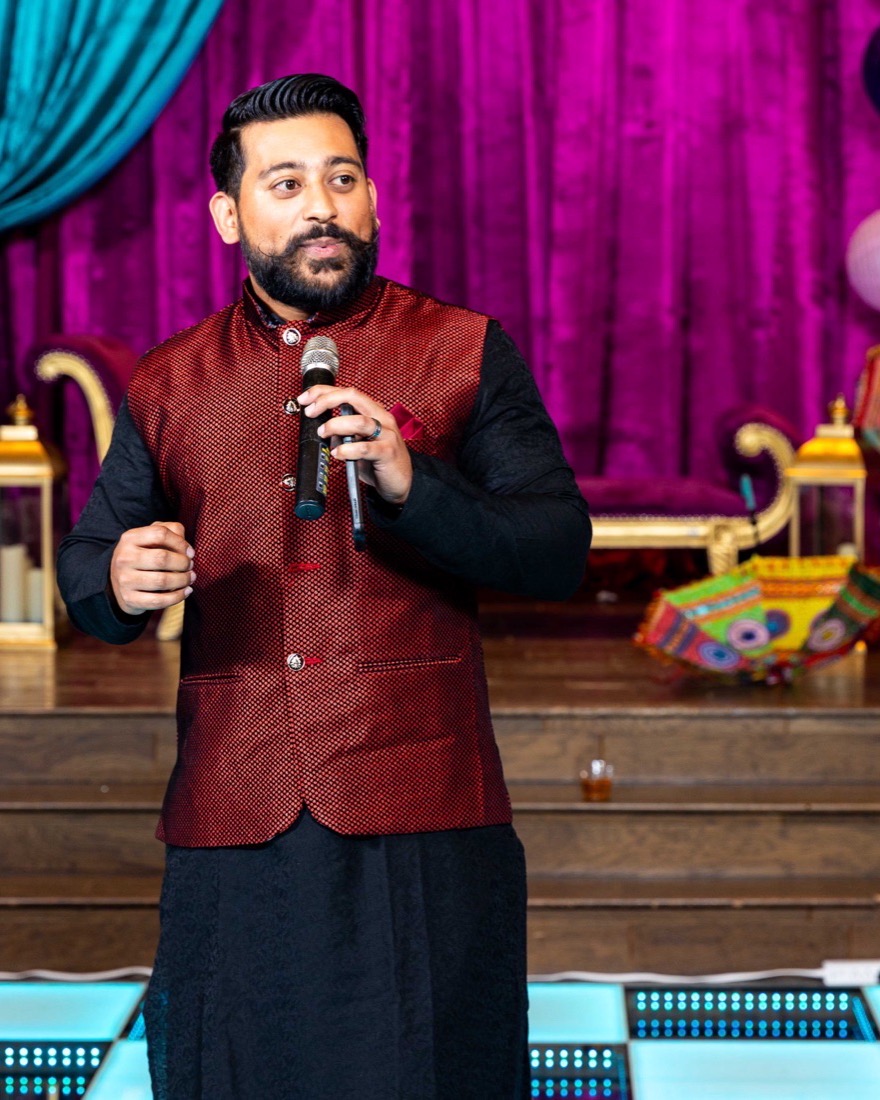 Anish holding a mic mid-toast at a sangeet, in a maroon bandi against a magenta stage
